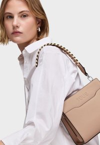 Młoda kobieta w białej koszuli trzyma beżową torebkę Tous z czarnym i beżowym plecionym paskiem na ramieniu na jednolitym tle.
