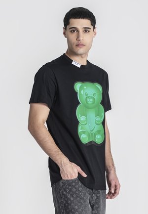 Jeune homme aux cheveux noirs courts portant un T-shirt noir avec un grand dessin d'ourson en gomme vert, debout avec une main dans la poche.