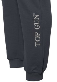 Dunkelgraue Sweatpants mit elastischen Bündchen und gesticktem "TOP GUN"-Text in Silber am linken Bein. Weiches Baumwollmaterial.