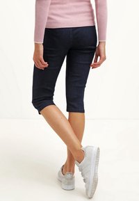 Mujer vistiendo pantalones cortos de mezclilla oscuros hasta la rodilla, una blusa de manga larga rosa y zapatillas blancas, de pie con las piernas cruzadas mirando hacia un lado.