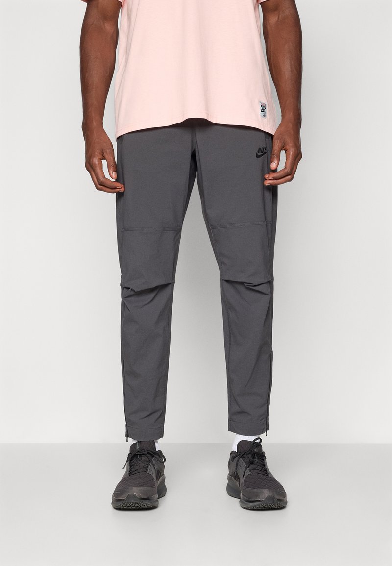 Szare spodnie sportowe Nike z tapered legs, elastycznym pasem i zapinanymi na zamek mankietami, w połączeniu z jasnoróżową koszulką i czarnymi trampkami.