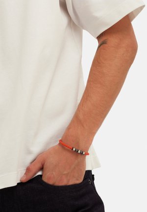 Geflochtenes orangenes Armband mit metallischen Akzenten, das zwei schwarze und silberne Perlen enthält, getragen an einer Hand gegen ein schlichtes weißes Hemd.