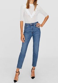 Eine Frau trägt eine weiße durchsichtige Bluse mit Punkten und Rüschen, hochgeschnittene blaue Jeans und schwarze spitze Ankle-Strap-Absätze.