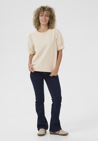 Top beige a maniche corte con maniche a sbuffo, abbinato a jeans svasati blu scuro e sneakers beige con dettagli chiari.