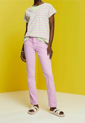 Persona con camiseta de rayas rosa y verde, pantalones rosa claro y sandalias beige, de pie contra un fondo amarillo.