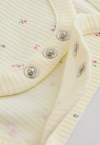 Zbliżenie kremowej prążkowanej tkaniny z drobnym różowym kwiatowym nadrukiem i srebrnymi zatrzaskami wzdłuż krawędzi otworu ubrania.