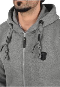 Sivi zip-up dukserica napravljena od mekanog materijala. Ima podstavljenu kapuljaču, crne akcentne detalje na vezicama i mali logo na grudima.