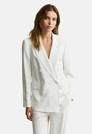 Frau mit hellbraunen Haaren, die einen weißen Doppelreiher-Blazer und passende weiße Hosen vor einem schlichten hellen Hintergrund trägt.