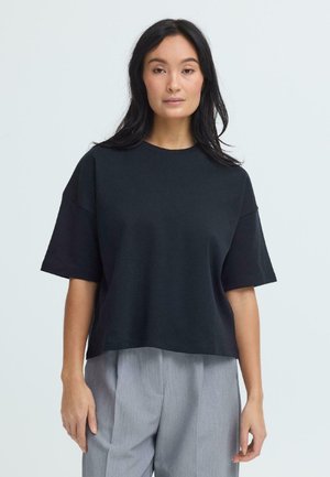 Vrouw met lang zwart haar, gekleed in een loszittende zwarte top met korte mouwen en lichtgrijze geplooide broek, staande tegen een effen achtergrond.