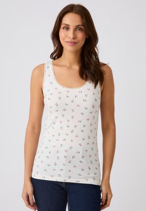 Woman wearing a white tank top with small red cherry prints and lace trim, paired with dark blue jeans, standing against a plain background.