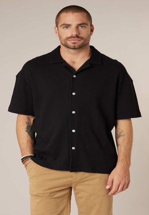 Man met kort haar en baard, gekleed in een zwart overhemd met korte mouwen en beige broek, staand met één hand in de zak.