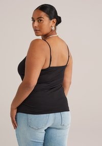 Frau mit dunklen Haaren im Dutt, die silberne Creolen, ein schwarzes Spaghettiträgershirt und eine hellblaue Jeans trägt, während sie über ihre Schulter schaut.