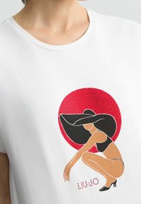 Weißes Baumwoll-T-Shirt mit einem grafischen Motiv einer Frau in einem schwarzen Hut und Badeanzug, umrahmt von Schwarz, vor einem roten Kreis mit einem gepunkteten Muster.