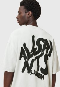 Joven con cabello rizado corto que lleva una camiseta blanca de gran tamaño con un gran texto ondulado en negro "ALSAINTS LONDON" en la parte posterior.