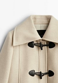 Beżowy płaszcz z wełny z szerokim kołnierzem, wyposażony w dwa czarne zapięcia na supełki i gładką fakturę. Dostosowane szwy podkreślają jego zarysowaną formę.