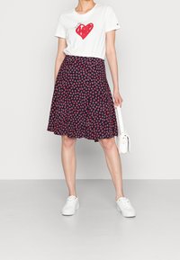 Camiseta blanca con un corazón rojo y texto, combinada con una falda azul marino que presenta un pequeño patrón de corazones rosa y rojo. Zapatillas blancas y bolso.