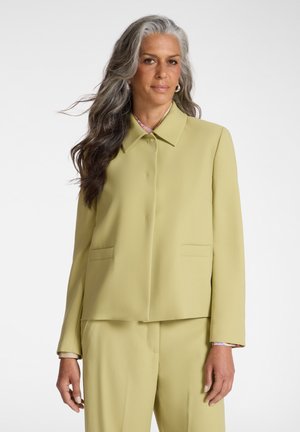 Mujer madura con cabello largo y gris, vistiendo una chaqueta de traje amarillo claro y pantalones a juego, de pie frente a un fondo blanco liso.
