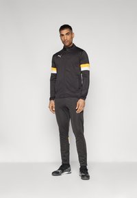 Μαύρο αθλητικό μπουφάν με πορτοκαλί και λευκές ρίγες στα μανίκια, στενά μαύρα παντελόνια και μαύρα αθλητικά παπούτσια με υφασμάτινη λεπτομέρεια.