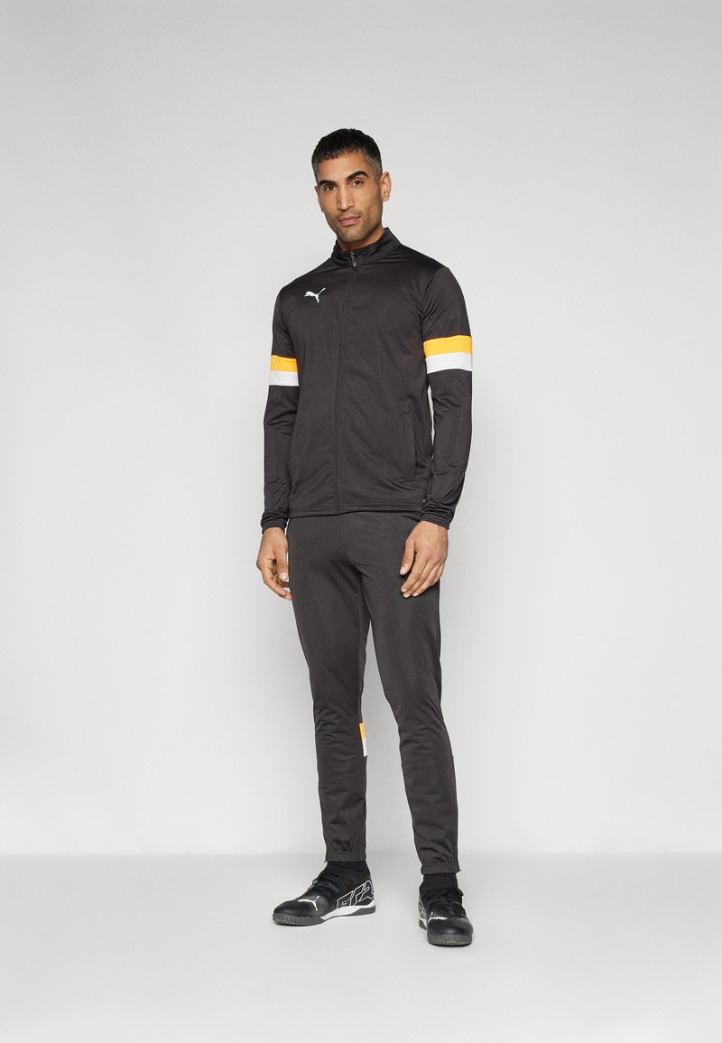 Μαύρο αθλητικό μπουφάν με πορτοκαλί και λευκές ρίγες στα μανίκια, στενά μαύρα παντελόνια και μαύρα αθλητικά παπούτσια με υφασμάτινη λεπτομέρεια.