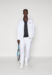 Hvit sportsjakke med teksturert mønster, kombinert med en svart t-skjorte og hvite bukser, holder en tennisracket. Har logo detaljer.