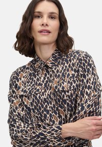 Granatowa koszula zapinana na guziki z wzorem zwierzęcym, z dwiema kieszeniami na przodzie i kołnierzykiem. Materiał jest gładki i lekki.