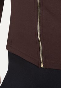 Haut marron avec fermeture éclair dorée. Le tissu semble extensible. Détails de couture visibles le long des bords. Tissu noir en dessous.