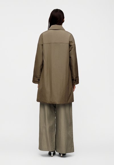 Hosszú sötét hajú nő háttal áll, barna hosszú kabátot, széles szárú nadrágot és fekete magassarkút visel, egyszínű világos háttér előtt.