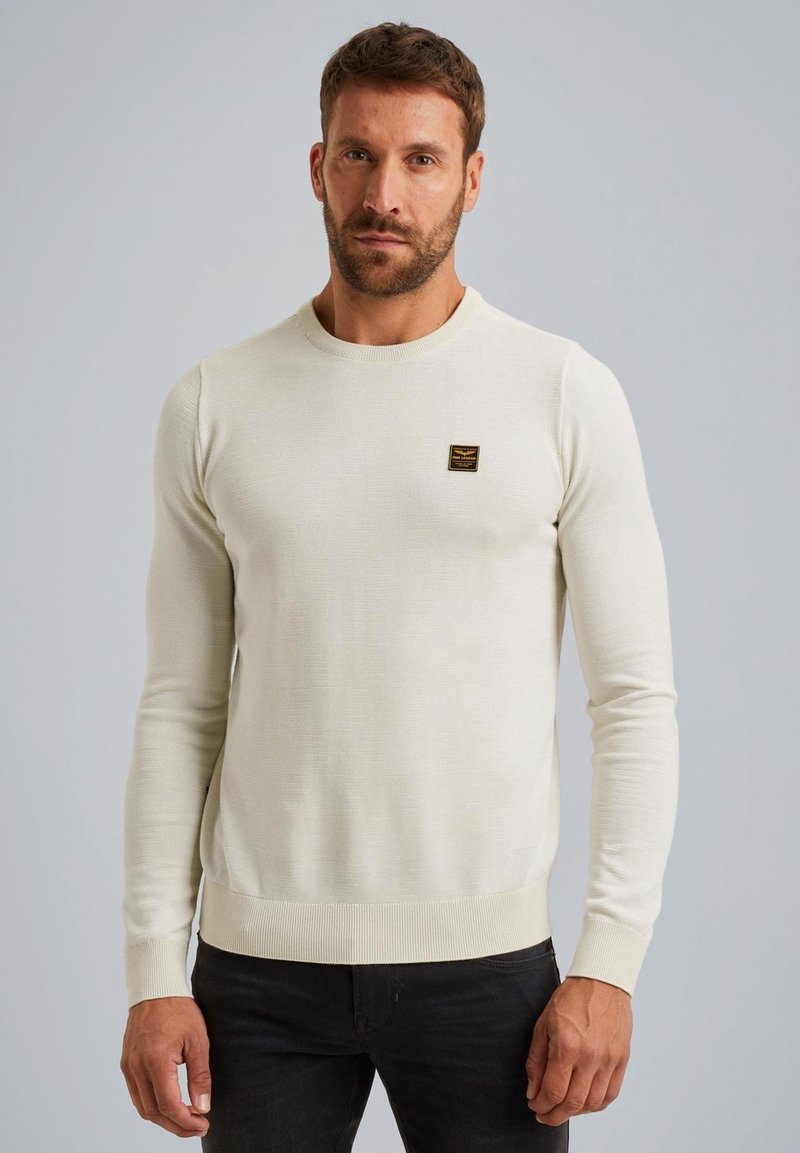 Maglione color crema realizzato in tessuto a maglia, con collo rotondo e polsini a coste. Include un piccolo etichetta nera sul petto.