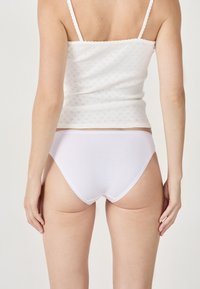 Accappatoio in cotone bianco con taglio bikini classico, caratterizzato da bordi lisci e una consistenza morbida. Indossato con una canotta bianca a fantasia.