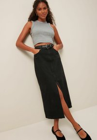 Melnas denima maksi svārki ar priekšēju iegriezumu, saskaņoti ar pelēku bez piedurknēm crop top un melnu jostu, valkāti ar melnām kurpēm.