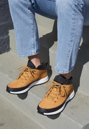 Person iført lyseblå jeans og brune vandresko med sorte og hvide såler, sidder på grå betonstrin.