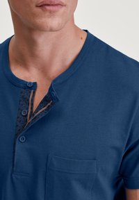 Korte mouwen blauwe shirt van zacht materiaal, voorzien van een knopenlijn bij de hals met een patroon aan de binnenkant en een borstzak aan de linkerkant.