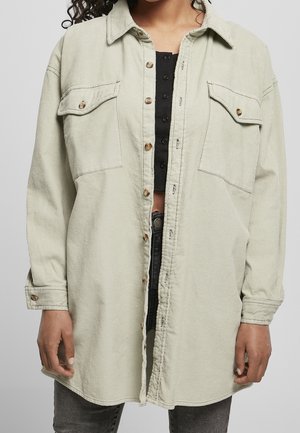 Femme portant une chemise en velours côtelé beige clair oversize par-dessus un crop top noir boutonné et un jean gris foncé sur un fond gris uni.