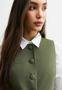 Chaleco verde oliva con tres botones redondos, usado sobre una camisa blanca con cuello clásico. La tela parece texturizada y estructurada.