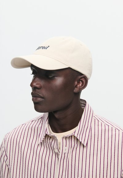 Šviesiai ruda medvilninė kepurė su kreiviu kraštu ir išsiuvinėtu užrašu. Dėvima su dryžuota marškinėliais, turinčiais purpurines ir baltas juostas.