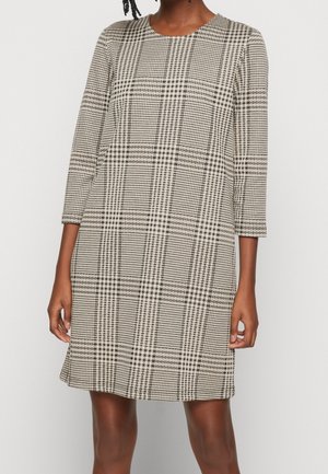 Frau trägt kniehohes, beige-schwarz kariertes Kleid mit dreiviertel Ärmeln und rundem Ausschnitt, steht mit entspannten Händen an den Seiten.