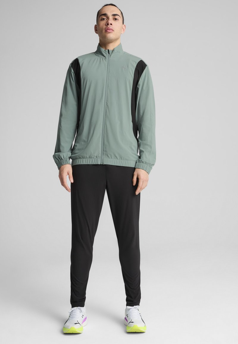 Leichte, mintgrüne Jacke mit schwarzen Seitenteilen, Reißverschluss vorn und elastischen Bündchen, kombiniert mit schwarzen Trainingshosen und weißen Sneakers.