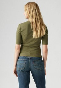 Olivgrön tajt topp med korta ärmar, parat med blå denimjeans med bakfickor och en röd etikett på högra fickan.