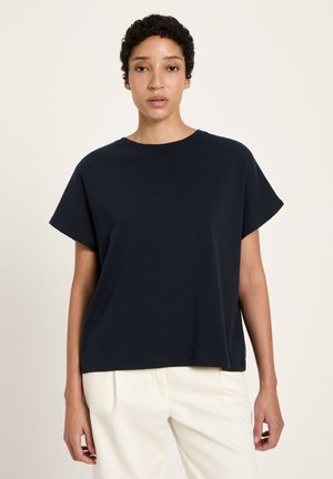 Frau trägt ein locker sitzendes schwarzes Kurzarmoberteil und hellfarbene Hosen, steht vor einem schlichten Hintergrund.