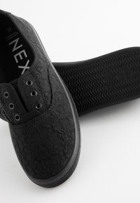 Sorte slip-on sko med blondermønster, gummisål og cirkulær tekstur. Har huller til snørebånd og polstret krave for komfort.