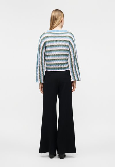 Femme vue de dos portant un pull en maille rayé bleu, vert et blanc avec des manches larges et un pantalon noir à jambes larges.