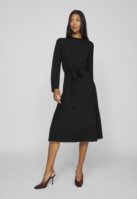 Black knitted dress with long sleeves, a belt at the waist, and a pleated skirt. Worn with pointed brown heels. Smooth texture, solid color.