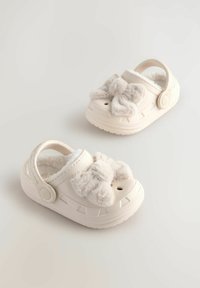 Sabots blancs pour enfants avec un détail de nœud en peluche, une tige texturée, une doublure douce et une sangle arrière. Conçus avec des trous de ventilation et une semelle antidérapante.