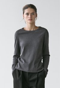 Femme aux cheveux tirés en arrière, portant un chemisier gris foncé à manches longues et un pantalon noir ample, les mains dans les poches, sur un fond clair uni.