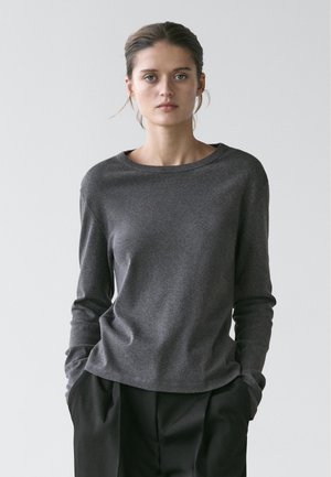 Kvinde med trukket hår tilbage iført en mørkegrå langærmet skjorte og løstsiddende sorte bukser med hænderne i lommerne mod en ensartet lys baggrund.