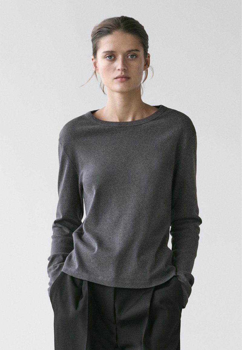 Femme aux cheveux tirés en arrière, portant un chemisier gris foncé à manches longues et un pantalon noir ample, les mains dans les poches, sur un fond clair uni.