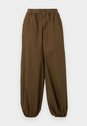 Løse brune bukser med elastiske mansjetter og linning med to knapper og sidelommer, vist på ensfarget bakgrunn.