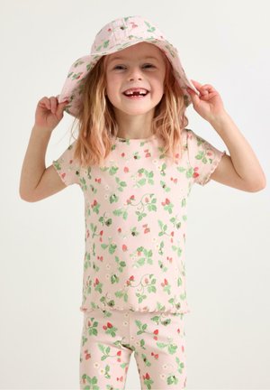 Glad ung pige iført lyserød blomsterhat, matchende skjorte og bukser, smilende med manglende fortænder mod en ensfarvet baggrund.