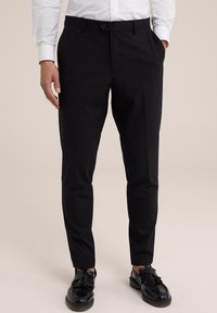 Černé kalhoty na míru s hladkým materiálem, slim fit, postranními kapsami a předním zipem. Zvýrazněny černými koženými botami s třásněmi.
