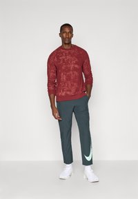 Mennenes antrekk med en teksturert burgunder skjorte med lange ermer og mørkegrønne bukser med en lys grønn logo. Hvite sneakers fullfører stilen.